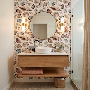 May include: A bathroom vanity with a light wood finish, a white vessel sink, and a matte black faucet. Above the vanity is a round mirror flanked by two cylindrical wall sconces. The wall features wallpaper with a repeating seashell pattern.