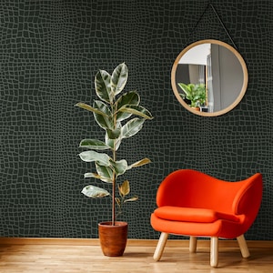 May include: A dark green crocodile-textured wallpaper covers the wall. A potted plant with large green and white leaves stands in front of the wall. An orange chair with wooden legs sits to the right. A round mirror hangs on the wall.