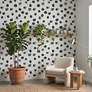May include: A room featuring white wallpaper with a black watercolor dot pattern, a large fiddle leaf fig in a terracotta pot, a wooden floating shelf with small potted plants, a cream-colored armchair, and a gold floor lamp next to a side table.
