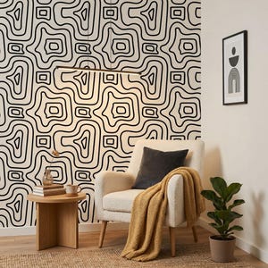 Pu&ograve; includere: Una poltrona color crema con un cuscino grigio scuro e una coperta in maglia giallo senape. Accanto, un tavolino rotondo in legno sostiene un piccolo vaso e una tazza. La parete presenta una carta da parati con un motivo a linee astratte.