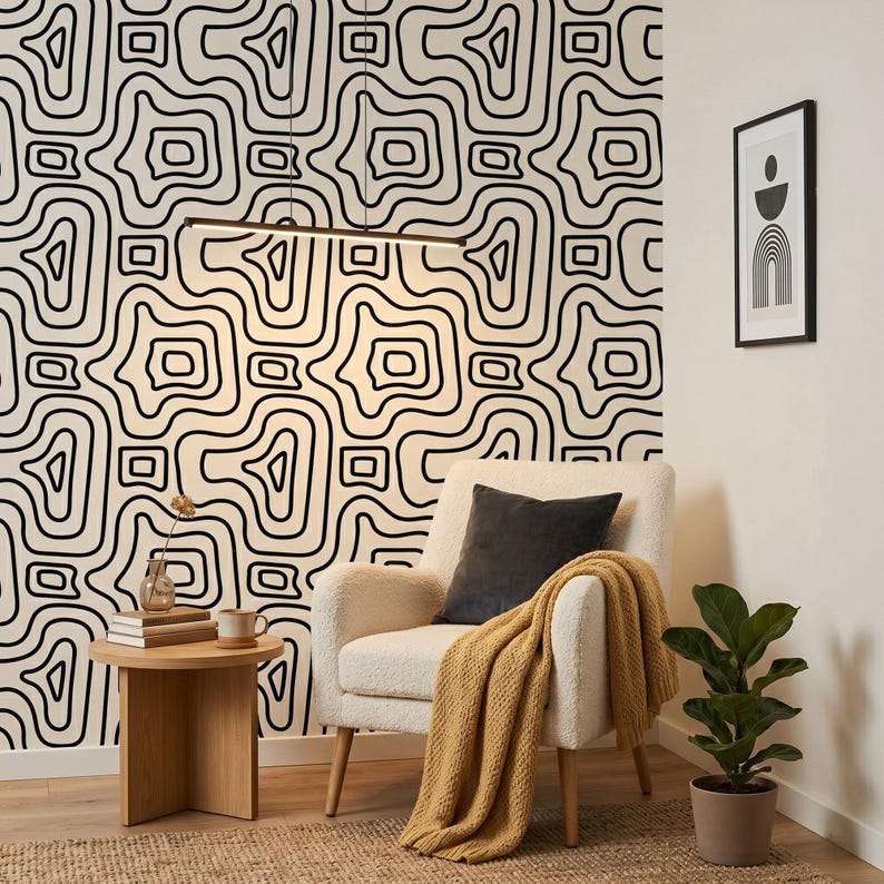 Pu&ograve; includere: Una poltrona color crema con un cuscino grigio scuro e una coperta in maglia giallo senape. Accanto, un tavolino rotondo in legno sostiene un piccolo vaso e una tazza. La parete presenta una carta da parati con un motivo a linee astratte.