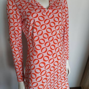 Puede incluir: Vestido de manga larga con cuello en V. El vestido presenta un patrón repetitivo de círculos naranjas sobre un fondo rosa claro. El vestido se exhibe en un maniquí.