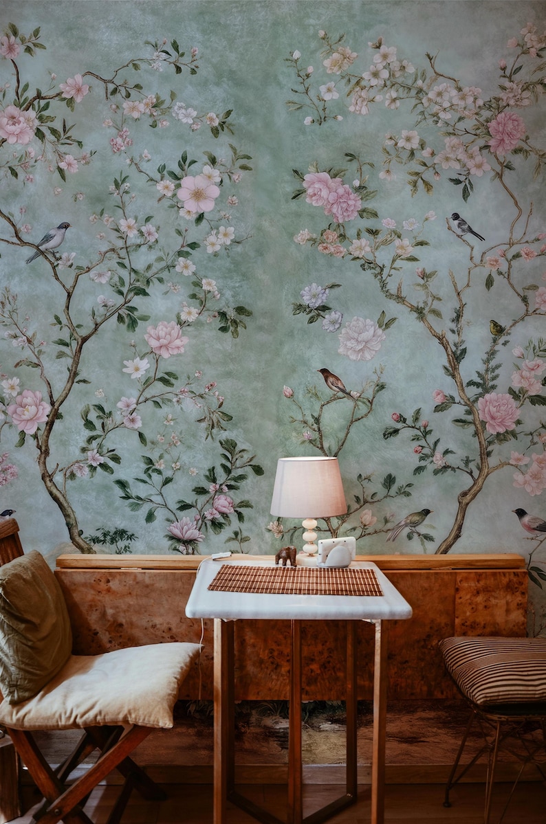 May include: A light green wallpaper with a floral pattern featuring pink flowers and small birds. A wooden table with a white tabletop and a lamp sits in front of the wallpaper.