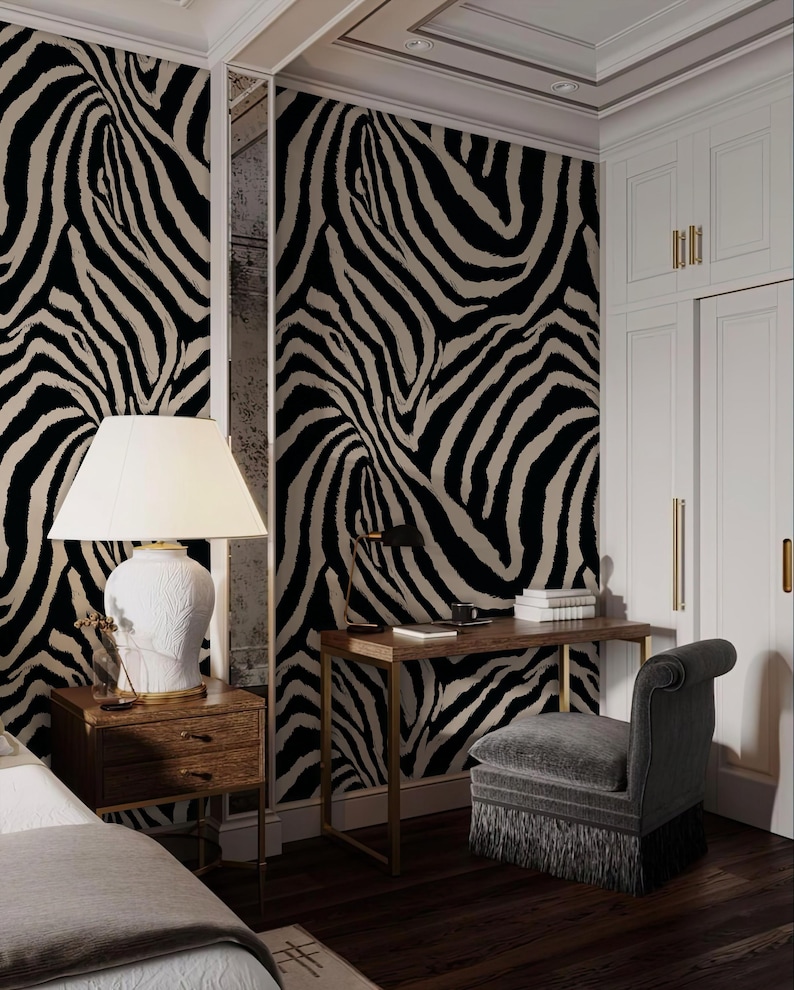 May include: A bedroom with zebra print wallpaper, a wooden desk with a gold frame, and a gray upholstered chair. A white lamp sits on a wooden nightstand. The room has white cabinets and a dark wood floor.