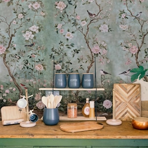 May include: A kitchen counter with a green cabinet, a wooden cutting board, a black metal stand with three jars labeled "tea", "coffee", and "sugar", and a floral wallpaper with pink flowers and birds.