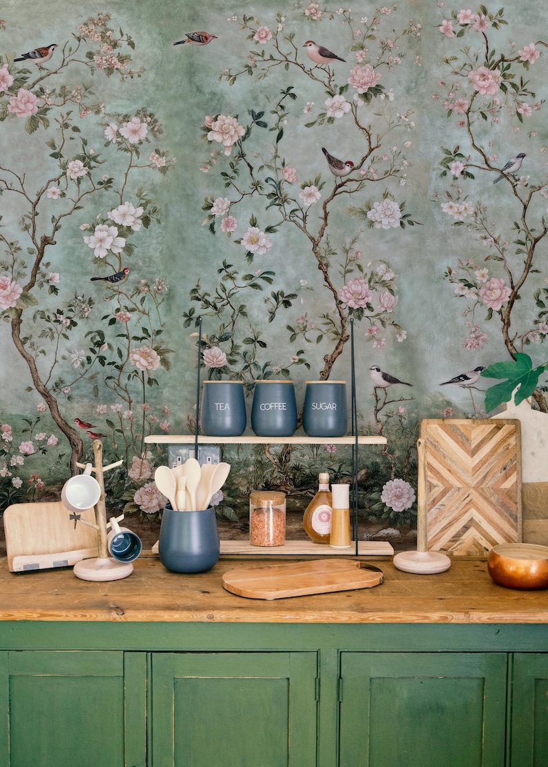 May include: A kitchen counter with a green cabinet, a wooden cutting board, a black metal stand with three jars labeled "tea", "coffee", and "sugar", and a floral wallpaper with pink flowers and birds.