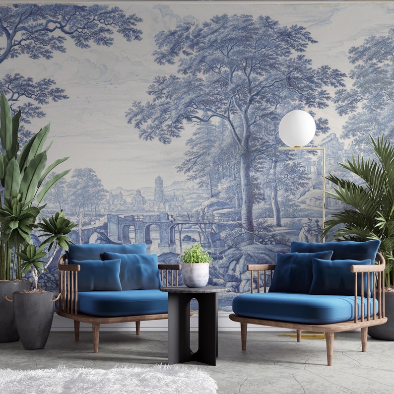 Puede incluir: Dos sillones de terciopelo azul con marcos de madera y una mesa auxiliar negra frente a un papel tapiz toile azul y blanco que representa un paisaje con un puente y &aacute;rboles.