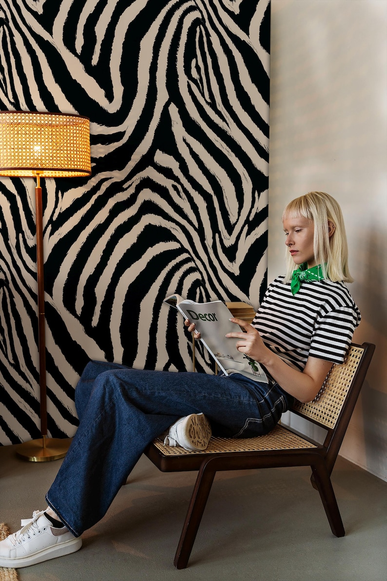 May include: A woman sits in a wooden chair, reading a magazine with the word "Decor" visible. She wears a striped shirt and jeans. The background features a zebra-print wall and a woven lamp.