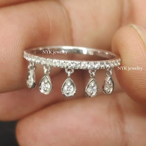 May include: A silver ring with a row of small, clear stones and six dangling teardrop-shaped gems. The ring is held in a hand against a dark background. The text "NYK jewelry" is visible on the ring.