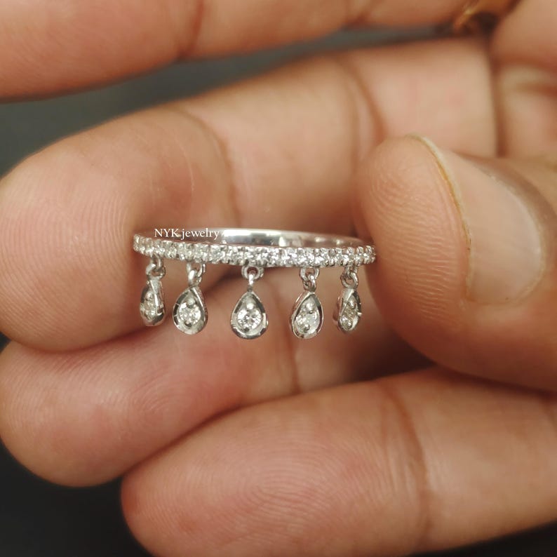 May include: A silver ring with a row of small, clear stones along the top. The ring has six dangling teardrop-shaped pendants, each with a clear stone. The ring is held in a person's hand.