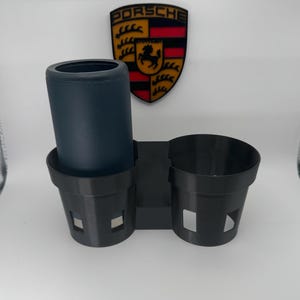 May include: A black car cup holder with two cup slots and a cylindrical holder. A dark blue tumbler is in the cylindrical holder. A Porsche emblem is in the background.