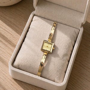 May include: A gold-toned wristwatch with a square face and a matching gold band. The watch is displayed in an open, beige velvet box. The watch face is a light cream color. The band has a textured design.