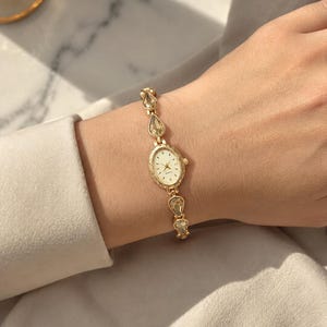May include: A gold-toned wristwatch with an oval face and a delicate band. The band features teardrop-shaped embellishments. The watch is worn on a wrist, showcasing its elegant design and classic appeal. The watch face has gold hands and markers.