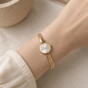 May include: A gold-toned wristwatch with a round face and a chain-link band. The watch face has a white background with black numerals. The watch is worn on a wrist, and the overall aesthetic is elegant and minimalist.