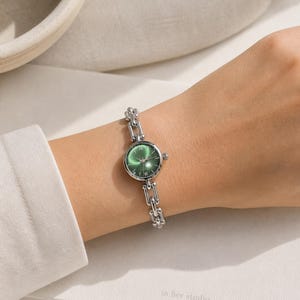 Könnte beinhalten: Eine silberne Armbanduhr mit grünem Zifferblatt und römischen Ziffern wird am Handgelenk getragen. Das Armband besteht aus kleinen, verbundenen Silbergliedern. Der Hintergrund zeigt eine weiße Oberfläche und den Text "in her studio 2022".