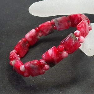 May include: A red and white gemstone bracelet. The bracelet features square-shaped beads with a marbled pattern, interspersed with smaller round beads. The color palette includes shades of red, white, and a hint of gray.