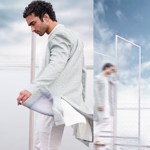 May include: A pale green embroidered jacket with a mandarin collar and long sleeves, worn with white trousers and a white shirt. The jacket has a delicate dotted design. The outfit is worn by a person with dark curly hair.