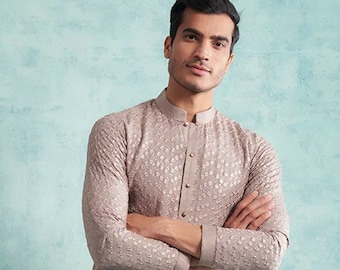 May include: A man is wearing a light grey kurta with a subtle pattern and matching trousers. He is standing against a blue background and wearing brown shoes.