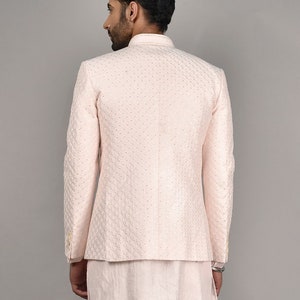 Puede incluir: Una chaqueta rosa claro con un patr&oacute;n acolchado y un cuello mandar&iacute;n. La chaqueta se lleva sobre una camisa rosa claro a juego y pantalones blancos.