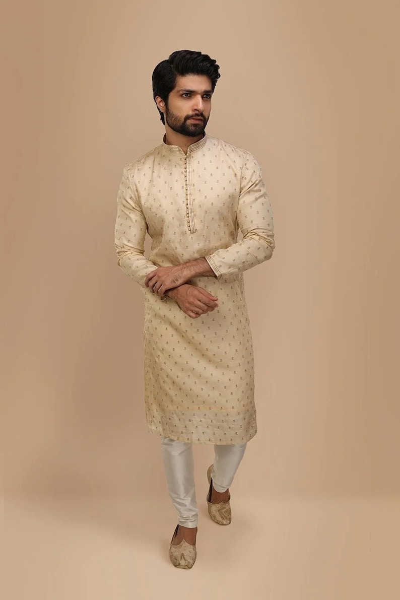 Op de afbeelding: Een cr&egrave;mekleurige, langarmige kurta met een knoopsluiting en een subtiel patroon. De kurta wordt gecombineerd met een witte broek en goudkleurige schoenen.