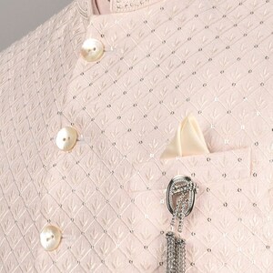 Puede incluir: Una camisa de botones rosa claro con un patr&oacute;n de diamante y botones de perla. La camisa tiene un bolsillo en el lado izquierdo con un alfiler decorativo de plata con una borla.