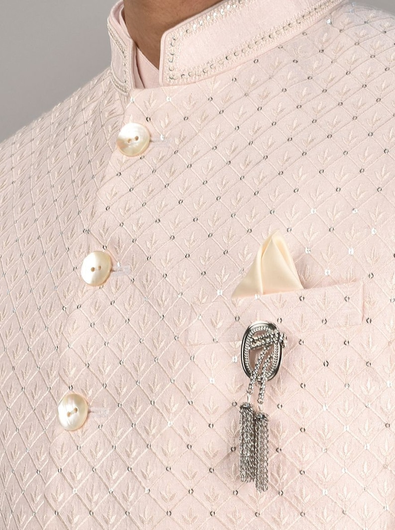 Puede incluir: Una camisa de botones rosa claro con un patr&oacute;n de diamante y botones de perla. La camisa tiene un bolsillo en el lado izquierdo con un alfiler decorativo de plata con una borla.