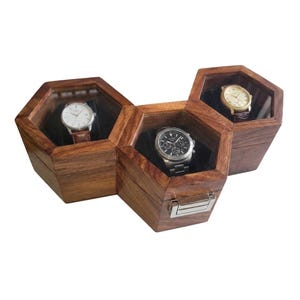 May include: Three connected hexagonal wooden watch boxes with clear glass lids, each containing a single wristwatch. The boxes feature a polished brown wood grain finish and silver-toned metal latches on the front, displaying three distinct timepieces.