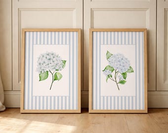May include: Two rectangular wall art prints in light wood frames. Each print features a watercolour illustration of a light blue hydrangea flower with green leaves, centred on a white background surrounded by a light blue and white vertical striped border.