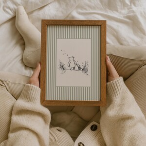 K&ouml;nnte beinhalten: Ein gerahmter Druck mit einer Schwarz-Wei&szlig;-Illustration eines B&auml;ren und eines kleinen Tieres, die zusammen gehen. Der Druck hat einen gestreiften Hintergrund in Graut&ouml;nen und ist in einem Holzrahmen.