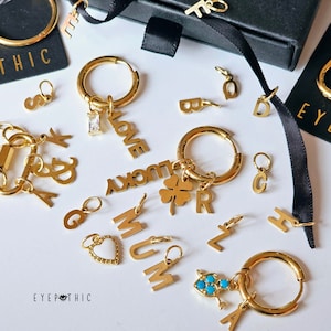 May include: A collection of gold-toned jewellery charms, including letters, words, and symbols. The charms are arranged on a white surface with a black background. The charms include the words "LOVE", "LUCKY", "MUM", and "EYEPHTHIC".