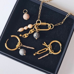 Op de afbeelding: Een gouden ketting met verschillende bedels, waaronder een marmerachtige steen, een parel en een gouden staaf. De ketting wordt gepresenteerd in een zwarte doos met twee gouden oorringen met bedels.