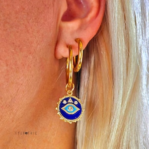 Op de afbeelding: Gouden creolen met een blauwe en witte hanger in de vorm van een boze oog. De hanger heeft een blauw oog met witte accenten en een gouden driehoek.