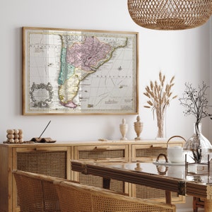 Può includere: Una mappa vintage del Sud America, incorniciata in una cornice di legno chiaro, è appesa a una parete bianca sopra un mobile in legno con ante in rattan intrecciato. La mappa è intitolata "TABULA GEOGRAPHICA PARS. CHILIS, PRETIA, MAG. LANICE" e mostra i paesi del Cile, del Perù e del Brasile.