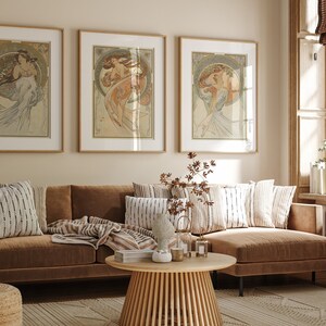 Puede incluir: Tres impresiones de arte enmarcadas de mujeres con vestidos fluidos, colgadas en una pared beige sobre un sof&aacute; seccional marr&oacute;n con almohadas y una manta. El sof&aacute; est&aacute; en una sala de estar con una mesa de caf&eacute; de madera y una alfombra tejida.