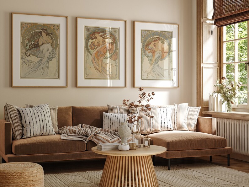 Puede incluir: Tres impresiones de arte enmarcadas de mujeres con vestidos fluidos, colgadas en una pared beige sobre un sof&aacute; seccional marr&oacute;n con almohadas y una manta. El sof&aacute; est&aacute; en una sala de estar con una mesa de caf&eacute; de madera y una alfombra tejida.