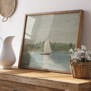 May include: A framed watercolor painting of a sailboat on a body of water with green hills in the background. The painting is on a wooden table with a white pitcher and a wicker basket of white flowers.