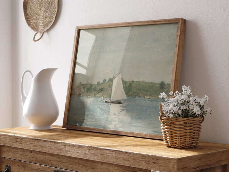 May include: A framed watercolor painting of a sailboat on a body of water with green hills in the background. The painting is on a wooden table with a white pitcher and a wicker basket of white flowers.