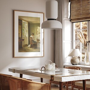 May include: A white pendant light with a textured shade hangs over a dining table with a glass top and a woven wicker chair. A framed print of a woman sitting at a table is on the wall behind the table.