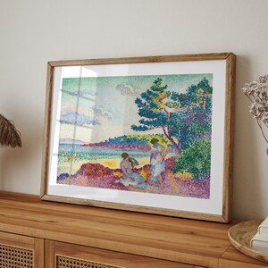 May include: A framed print of a pointillist painting depicting three figures on a beach. The figures are wearing white clothing and are standing under a large tree. The background is a blue sky with white clouds and a body of water.
