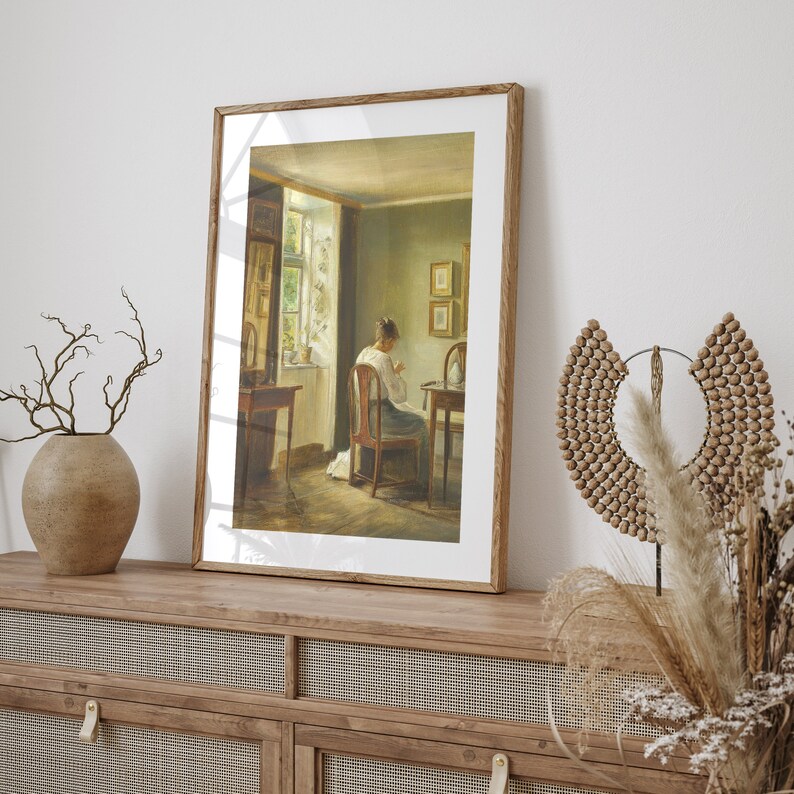 May include: A framed painting of a woman sitting in a chair in a room with a window. The woman is wearing a white dress and is working on something in her lap. The room is decorated with a few pieces of furniture and a window with a view of a garden.