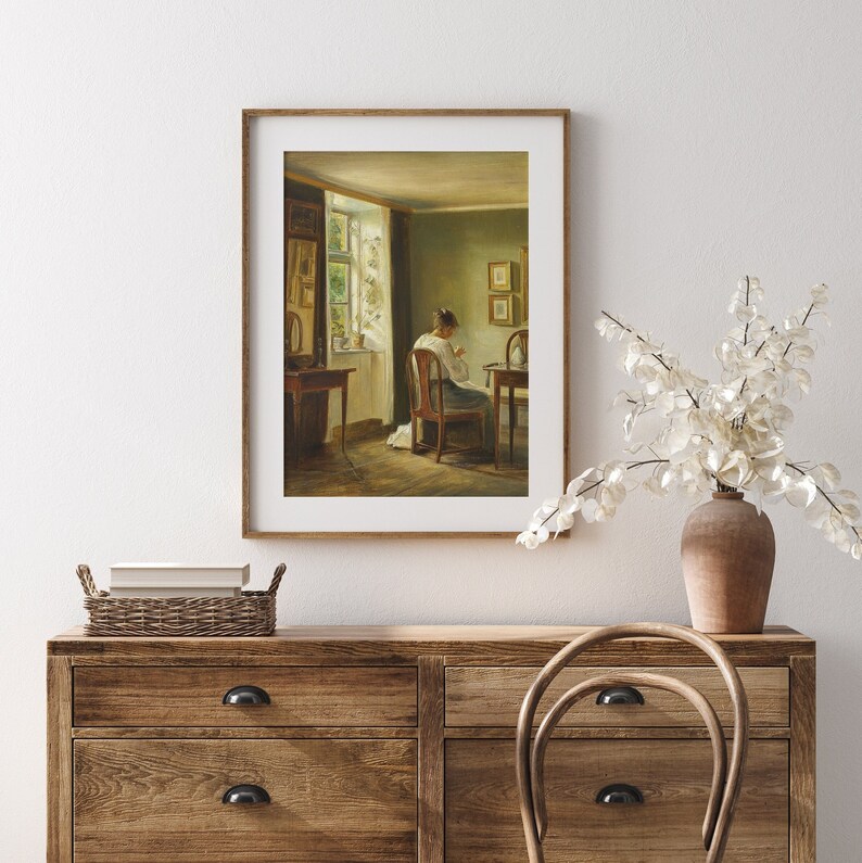 May include: A framed painting depicting a woman sitting in a chair in a room with a window. The woman is wearing a blue dress and is sewing. The room is decorated with a green wall and a wooden floor. The window is open and there is a view of a garden outside.