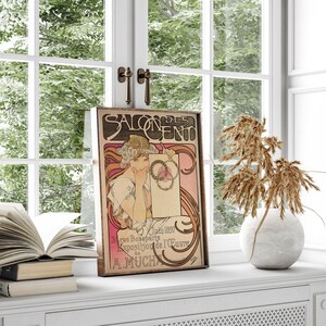 May include: A vintage poster for the Salon des Cent, featuring a woman in a white dress holding a wreath of flowers. The poster is in a wooden frame and has a pink and white background with the text "Salon des Cent" in black lettering. The text at the bottom of the poster reads "15 Juin 1897 31 rue Bonaparte Exposition de l'Oeuvre de A. Mucha."