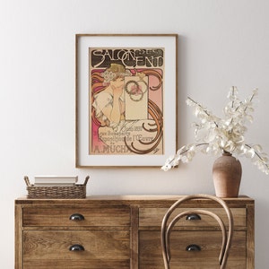 May include: A vintage poster advertising the Salon des Cent, an exhibition of artwork by Alphonse Mucha. The poster features a woman with flowing red hair, wearing a floral crown, holding a blank card with the text "Salon des Cent" in a decorative font.