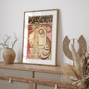 May include: A vintage poster for the Salon des Cent exhibition in Paris, France. The poster features a woman in a white dress with a floral crown, holding a scroll with the text "Salon des Cent". The poster is framed in a wooden frame.