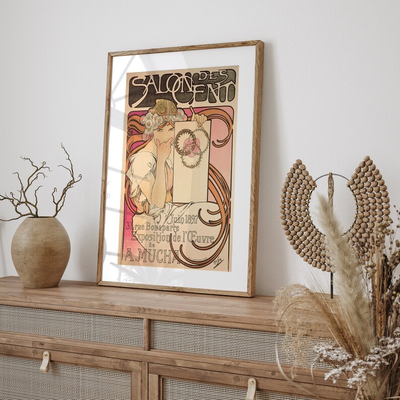 May include: A vintage poster for the Salon des Cent exhibition in Paris, France. The poster features a woman in a white dress with a floral crown, holding a scroll with the text "Salon des Cent". The poster is framed in a wooden frame.