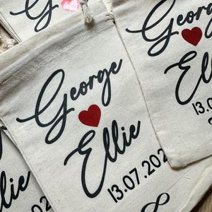 May include: White cotton drawstring bags with black calligraphy script reading "George & Ellie" with a red heart and the date "13.07.2022" printed on the front. The bags are ideal for wedding favours or gifts.