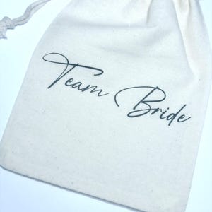 May include: A white cotton drawstring bag with the text "Team Bride" printed in black.