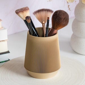 Puede incluir: Un conjunto de brochas de maquillaje en un recipiente beige acanalado. El recipiente se encuentra sobre una superficie de madera redonda y blanca, con una base de rodaja de madera natural. Un jarrón blanco con flores secas y libros en el fondo.