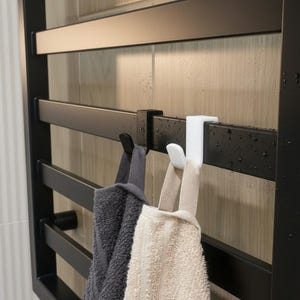 Può includere: Un termoarredo nero con due asciugamani appesi. Un asciugamano è grigio e l'altro beige. Il termoarredo è montato su una parete piastrellata. Gli asciugamani sono appesi a ganci bianchi.