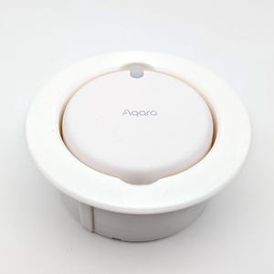 Puede incluir: Un dispositivo domótico blanco con la marca "Aqara" impresa en la parte superior. El dispositivo tiene un diseño circular con un centro empotrado y un anillo circundante. El dispositivo está sobre un fondo blanco.